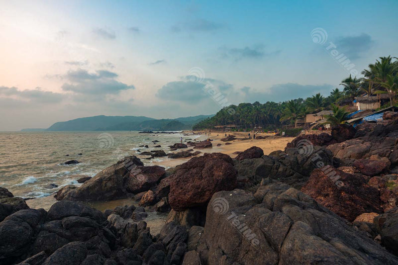 Experience the Serenity of Cola Beach, Goa - Small Waves Crashing on Rocky Cobbles as Water Recedes and Trees Line the Shoreline.